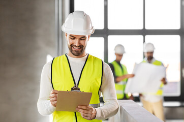 architecture, construction business and building concept - happy smiling male architect in helmet and safety west with clipboard working at office