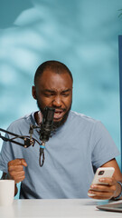 Cheerful vlogger recording video to give good news to audience for podcast. Blogger feeling happy while looking at smartphone and talking to subscribers using microphone for vlog.