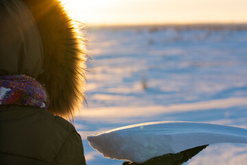 A girl in a hood with a small snowball on a sunset background. Back view. Natural frame. - 479732743