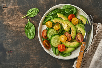 Avocado, red, yellow, black cherry tomato, spinach and cucumber fresh salad with spices pepper and olive oil in grey bowl old wooden table background. Healthy food concept. Top view.