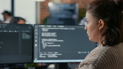 Over shoulder view of focused database designer writing code looking at multiple computer screens displaying algorithm. System engineer coding while colleagues doing teamwork in background.