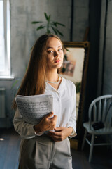 Vertical portrait of elegance young woman pianist holding in hands score with musical notes in dark classroom, looking at camera. Focused female musician student posing in room with vintage interior.