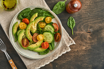 Avocado, red, yellow, black cherry tomato, spinach and cucumber fresh salad with spices pepper and olive oil in grey bowl old wooden table background. Healthy food concept. Top view.