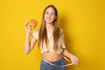Portrait of a happy young woman holding measuring tape around her waist and a croissant isolated over yellow background