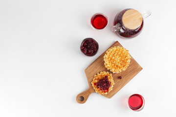 Thin waffles on a wooden board with berry jam on a white table, hibiscus tea. Tea party with sweets.
White background, top view, copy space.