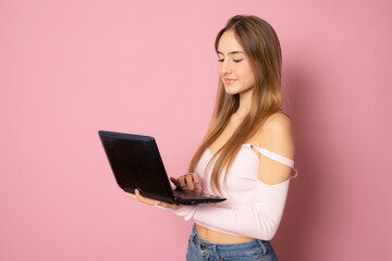 Beautiful girl with long hair using laptop computer standing isolated over pink background.