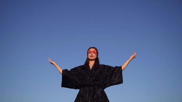 Brunette superwoman in black dress and red face mask spreading hands and looking up praying on clear blue sky background. Female power, protest, women rights, activism concept. 4k video footage