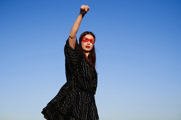 Portrait of strong supergirl in black dress and red face mask making fist pump, protesting for...