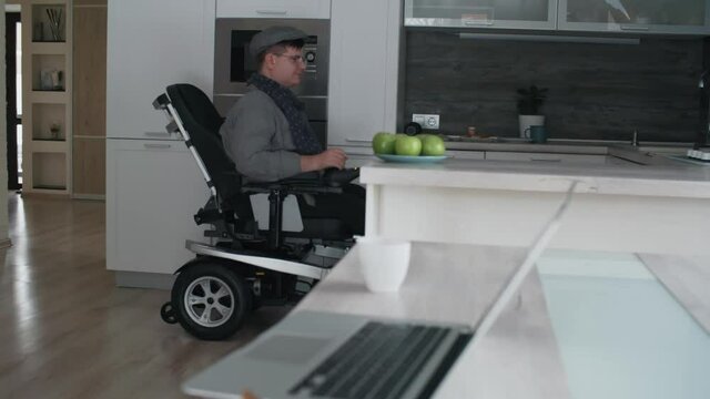 Low angle of Caucasian man in wheelchair wearing eyeglasses and peaked cap, entering kitchen, taking green apple from plate on cutting table