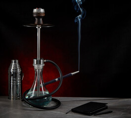 Beautiful modern hookah, from the mouthpiece of which smoke rises, stands on the table, surrounded by a metal protective cap and a diary, on a dark red background. Space for copying.