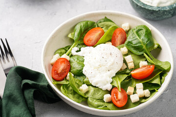 Salad bowl with fresh green spinach, cherry tomatoes, croutons, egg benedict and sour cream dressing on a grey stone surface