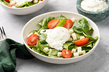 Salad bowl with fresh green spinach, cherry tomatoes, croutons, egg benedict and sour cream dressing on a grey stone surface