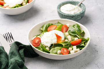 Salad bowl with fresh green spinach, cherry tomatoes, croutons, egg benedict and sour cream dressing on a grey stone surface