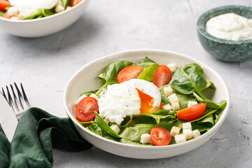 Salad bowl with fresh green spinach, cherry tomatoes, croutons, egg benedict and sour cream dressing on a grey stone surface