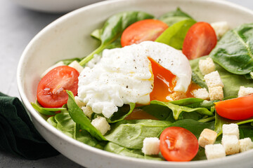 Salad bowl with fresh green spinach, cherry tomatoes, croutons, egg benedict and sour cream dressing on a grey stone surface