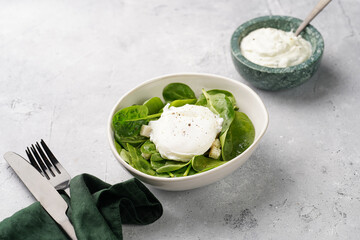 Small white salad bowl with fresh green spinach, egg benedict and sour cream dressing on a grey stone surface