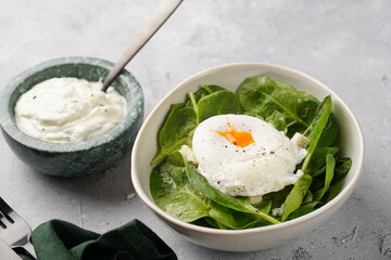 Small white salad bowl with fresh green spinach, egg benedict and sour cream dressing on a grey stone surface