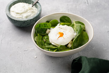 Small white salad bowl with fresh green spinach, egg benedict and sour cream dressing on a grey stone surface