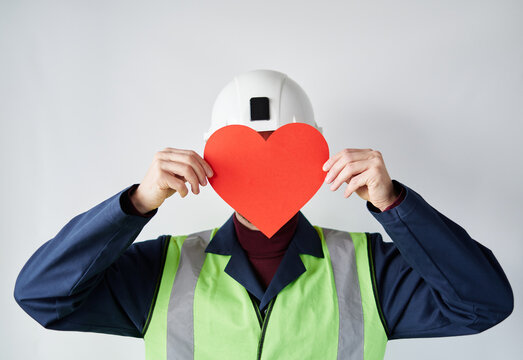 Portrait of bearded construction site worker male standing by the wall holding red heart shape. Worker with paper heart in front of the face. Valentine's Day concept. High quality image