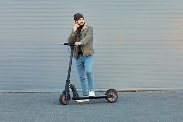 Man with electric scooter near wall. Ecological transportation concept. Stylish bearded male using phone, posing on e-scooter.