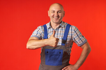 Bald man in overalls and a plaid shirt stands on a red background and shows a thumbs-up gesture....