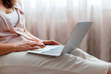 Naklejka premium Calm woman use laptop at cozy home interior. Close up female hands typing on keyboard, working using laptop or do online shopping at home on pink chair near window.