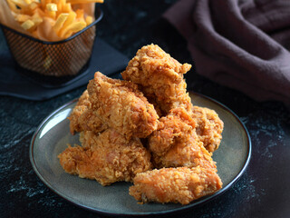 Spicy fried chicken wings, breaded on a ceramic plate. There are fries and a brown cotton towel in the background. Photo on a dark background. Low key. Minimalism. Selective focus.