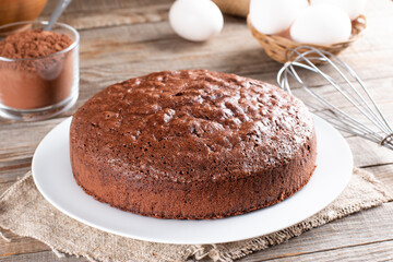 Fototapeta na wymiar Homemade round chocolate sponge cake or chiffon cake on plate so soft and delicious with ingredients: eggs, flour, cocoa, milk on wood table. Homemade bakery concept for background and wallpaper