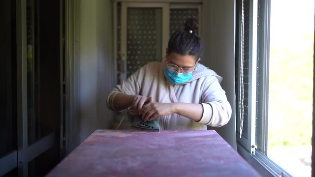 Chica joven guapa con mascarilla lijando un mueble para restaurarlo 
