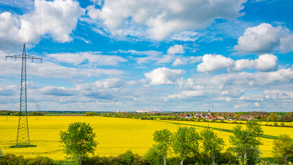 Panoramic view over beautiful farm countryside landscape with rapeseed yellow at blossom field, with a village at horizon in Germany, Spring, and high voltage power lines at blue sky and sunny day.