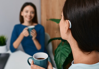Adult woman with a hearing impairment uses a hearing aid to communicate with her female friend during tea drinking at home. Hearing solutions - obrazy, fototapety, plakaty