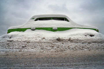 Old car covered with deep snow blocked in a large snowdrift on the street in cold winter day after...