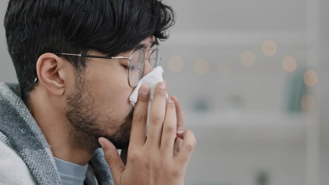 Close-up male face side view sick arabic hispanic man bearded guy with glasses wrapped in blanket suffers from runny nose sneezes allergy wipes snot with paper napkin respiratory disease infection