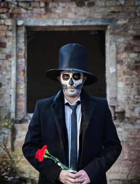 A man with skull make-up in the form of death in a tailcoat and a top hat near the ruins of an old building. The day of the Dead. Halloween. Red clove.