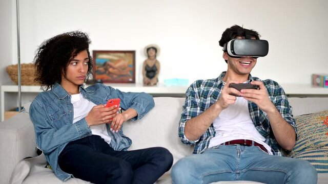 African American girl and mixed race guy spend leisure time together at home a sofa. Excited guy uses virtual reality glasses for video game, a bored girl sits next to him and looks at him indignantly