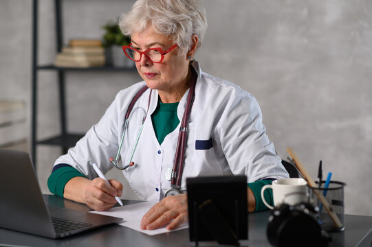 Middle age female doctor using laptop, writing notes with stethoscope on table. Healthcare concept. Medical doctor writing and taking notes