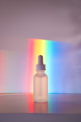 Face serum in matte glass bottle with a dropper in front of vibrant rainbow prism background. Front view