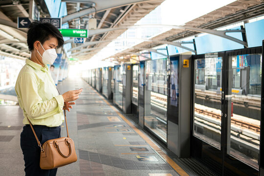 Asian female wearing medical face mask using mobile phone while waiting mass rapid train in empty subway platform during coronavirus outbreak situation.
