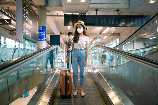 Asian female wearing medical face mask with travel suitcase at escalator in airport terminal. New normal concept of traveler awareness and prevention for omicron covid-19 situation. - Powered by Adobe