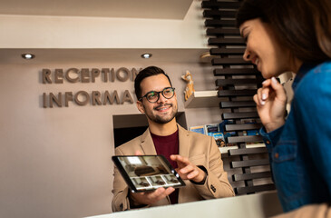 Smiling male receptionist behind the hotel counter showing guest available rooms on tablet.
