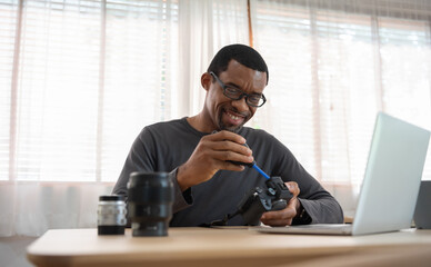 Fototapeta na wymiar Portrait of Smiling African American photographer man cleaning digital camera with a Rubber air blower, Happy Black Freelancer in home studio