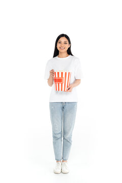 full length view of joyful asian woman with large bucket of popcorn looking at camera on white.