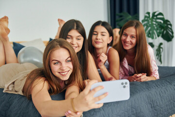 Laying down on bed with smartphone. Group of happy women that is at a bachelorette party