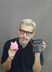 Wise middle age men with gray hair and beard in eyeglasses holding pink piggy bank and calculator on gray background. Saving money concept.