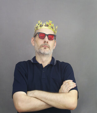 Middle age men with gray hair and beard in red eyeglasses glasses and golden king crown on head on gray background