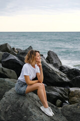 A young attractive Caucasian blonde woman of 35 years sits on the rocks of sea coast. Emerald-colored water, waves splashing on the stones. The woman meditates and relaxes.