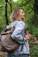 A young Caucasian woman walks along a forest mountain road. Casual blue denim clothes and backpack. Travel, healthy lifestyle and freedom.