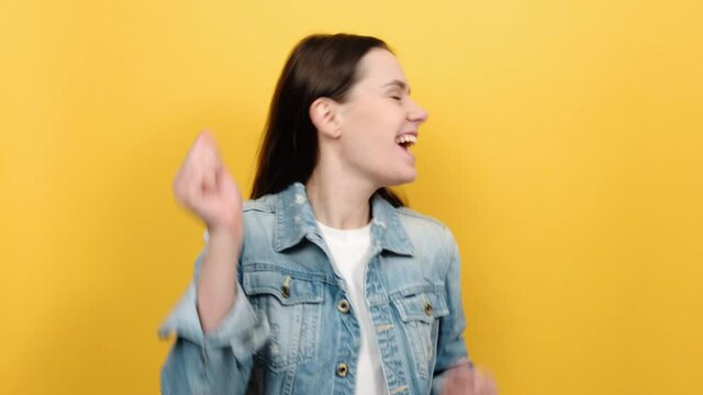 Euphoric thrilled young woman in denim jacket sincerely laughing and rejoicing, celebrating her lottery victory, success, posing isolated over yellow color background studio. People emotion concept