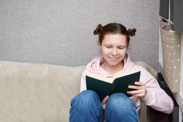 Young happy woman sitting with her legs bent under her on the couch in a room in her home, smiling broadly, resting on a day off spends time usefully reads a book, copy space.