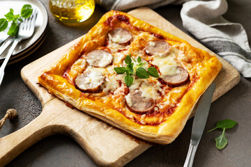 Homemade puff pastry pizza with salami, mozzarella, tomatoes and basil on the kitchen table. Delicious layered pie with sausage and cheese on a dark culinary background closeup